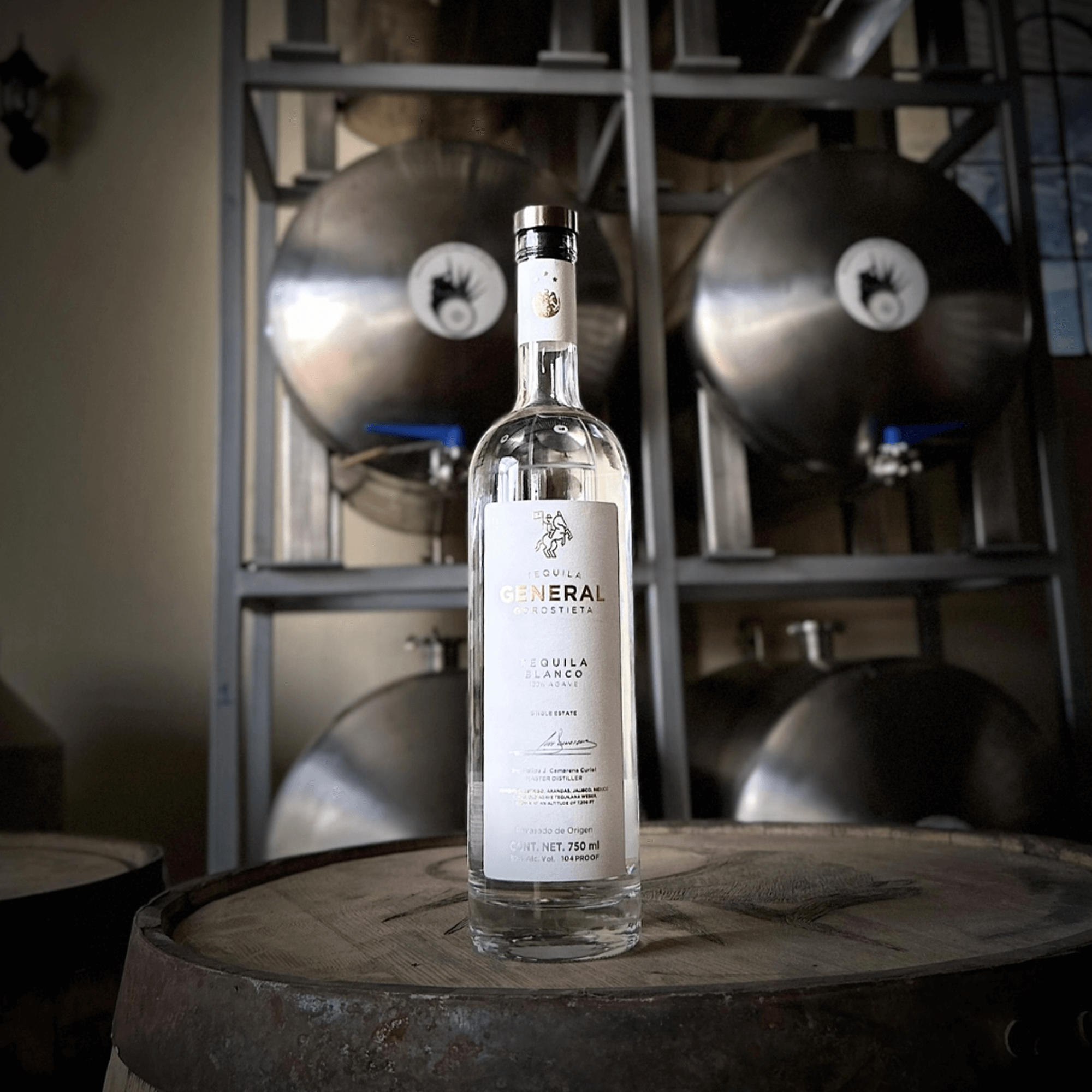Bottle of General Gorostieta High Proof Blanco Tequila on a wooden barrel, with fermentation tanks in background.