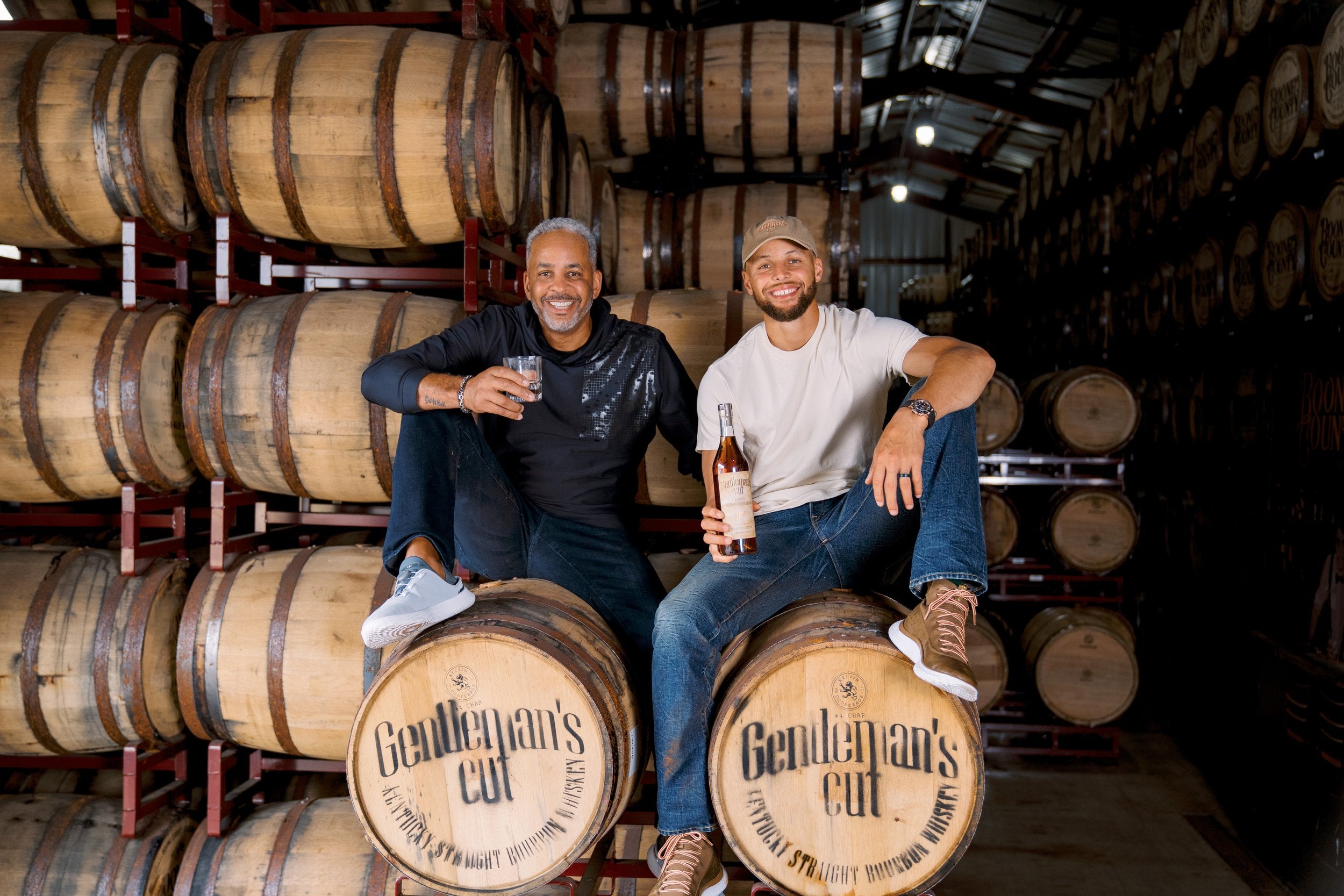 Two men enjoying Gentleman's Cut bourbon in a distillery surrounded by barrels, celebrating a special legacy.
