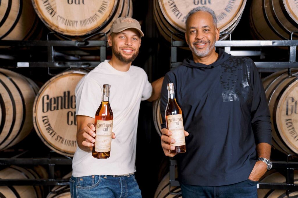 Father and son holding Gentleman's Cut bourbon bottles in front of whiskey barrels.