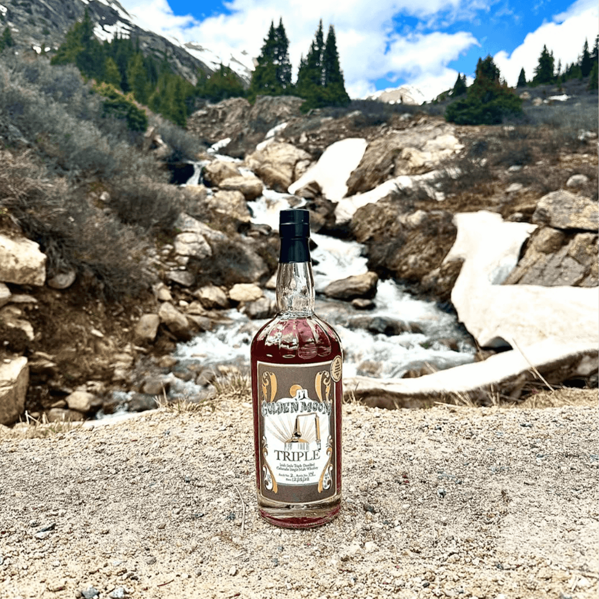 Golden Moon Triple Colorado Single Malt Whiskey bottle in a scenic mountain setting near a flowing stream.