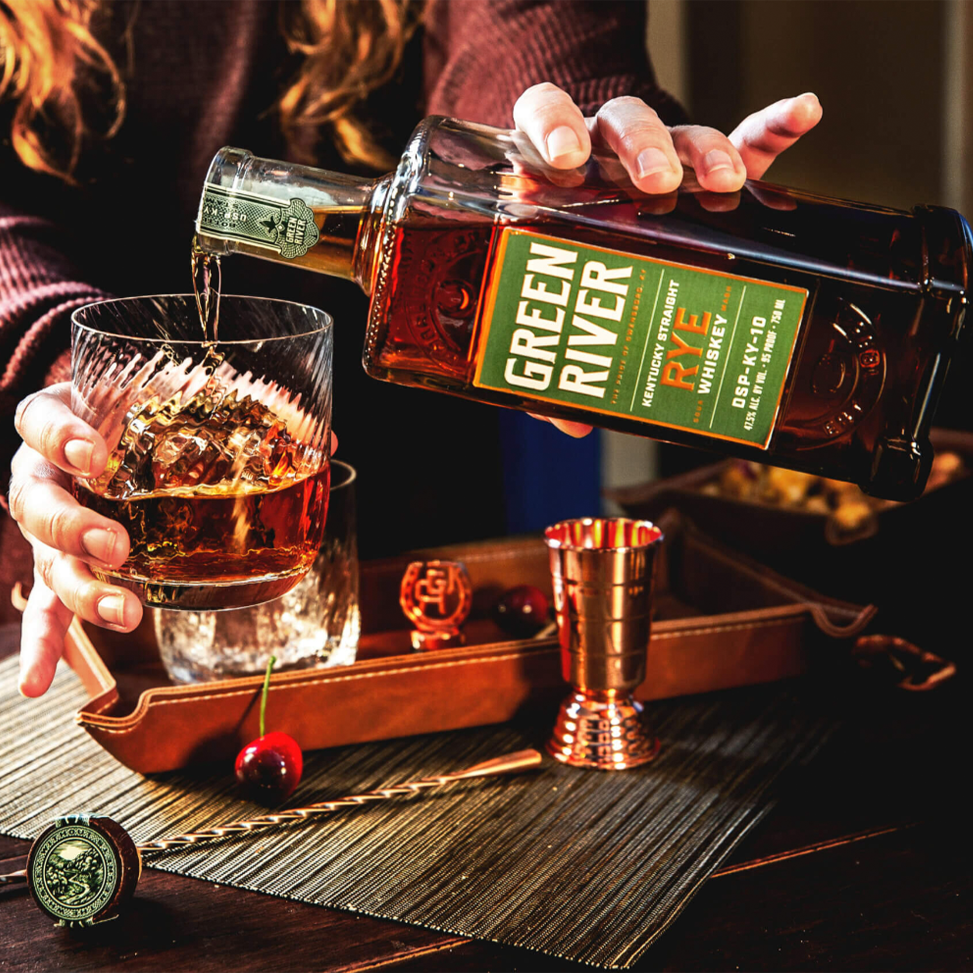 Pouring Green River Kentucky Straight Rye Whiskey into a glass with a copper jigger and cherries.