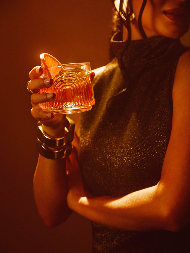 Woman in a glittery outfit holding a glass with a grapefruit slice, showcasing a cocktail inspired by 1800 Guachi Monton Añejo Tequila.
