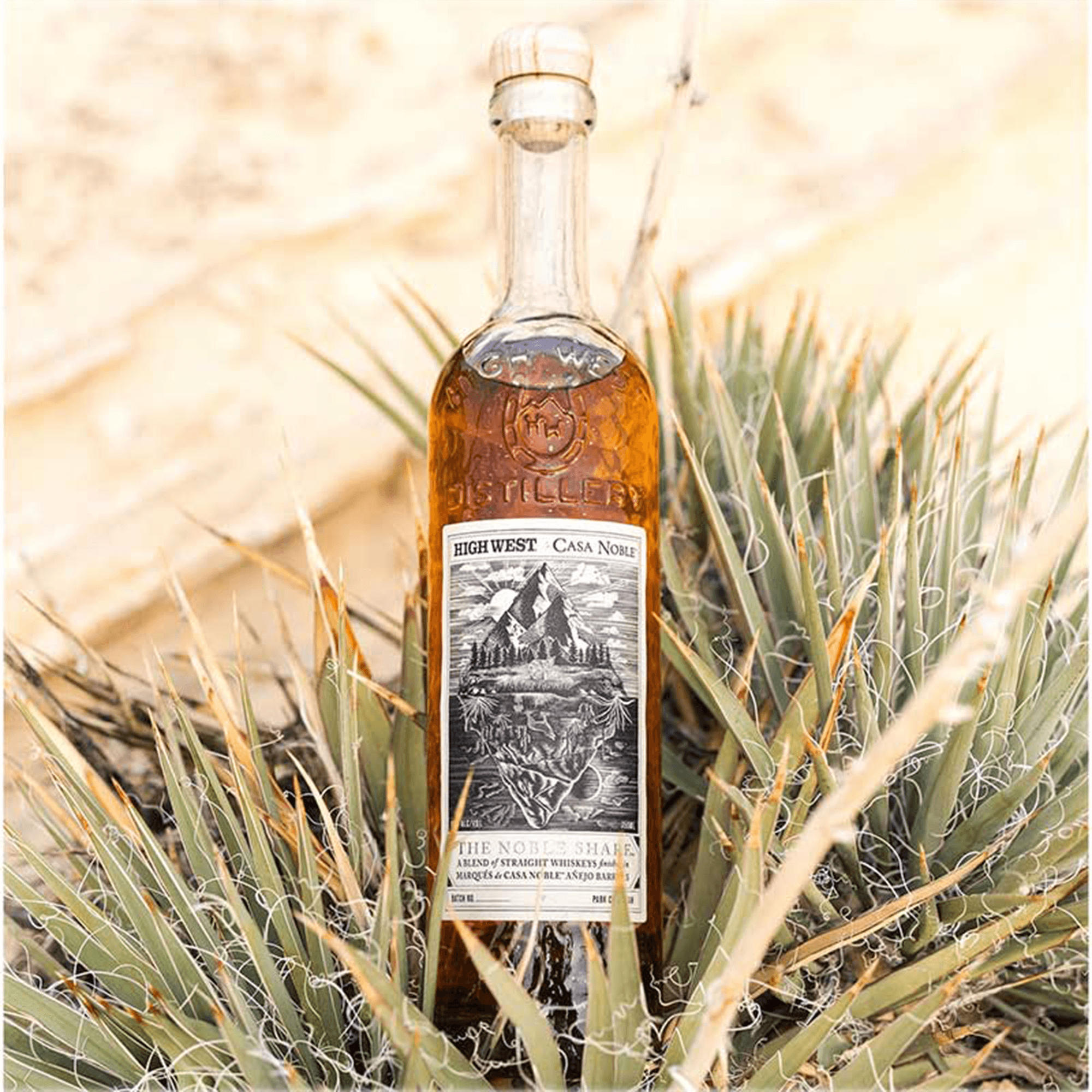 High West x Casa Noble The Noble Share whiskey bottle surrounded by agave plants in a desert setting.