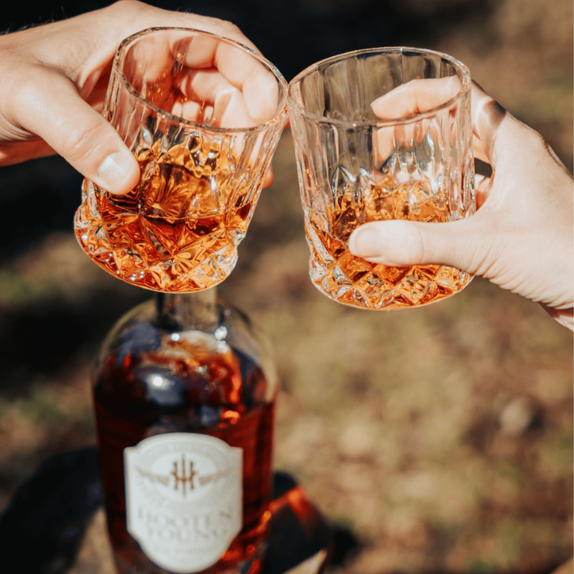 Two hands toasting with glasses of Hooten Young Rye Whiskey against a bottle outdoors.