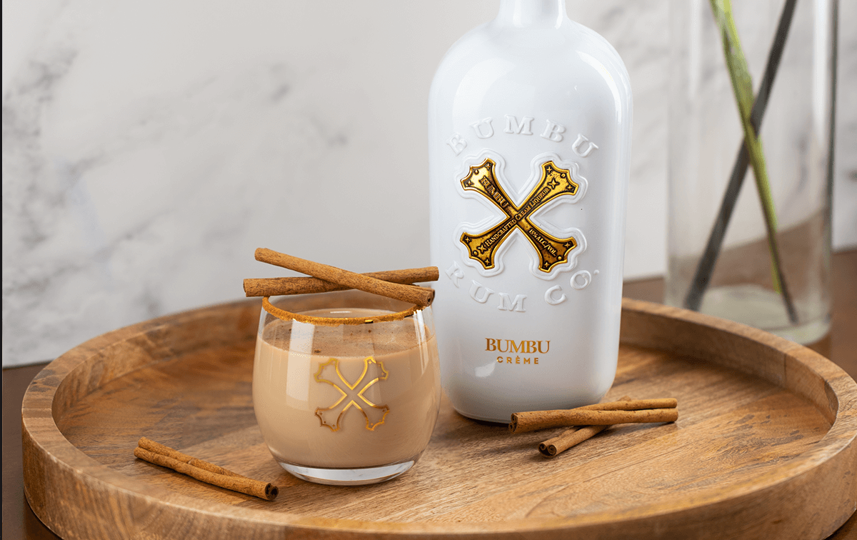 Bumbu Crème Rum bottle and glass with creamy rum and cinnamon sticks on a wooden tray.