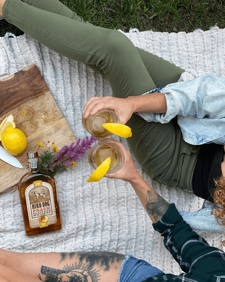 Two people enjoying Bird Dog Peach Whiskey cocktails outdoors with fresh lemons and flowers on a picnic blanket.