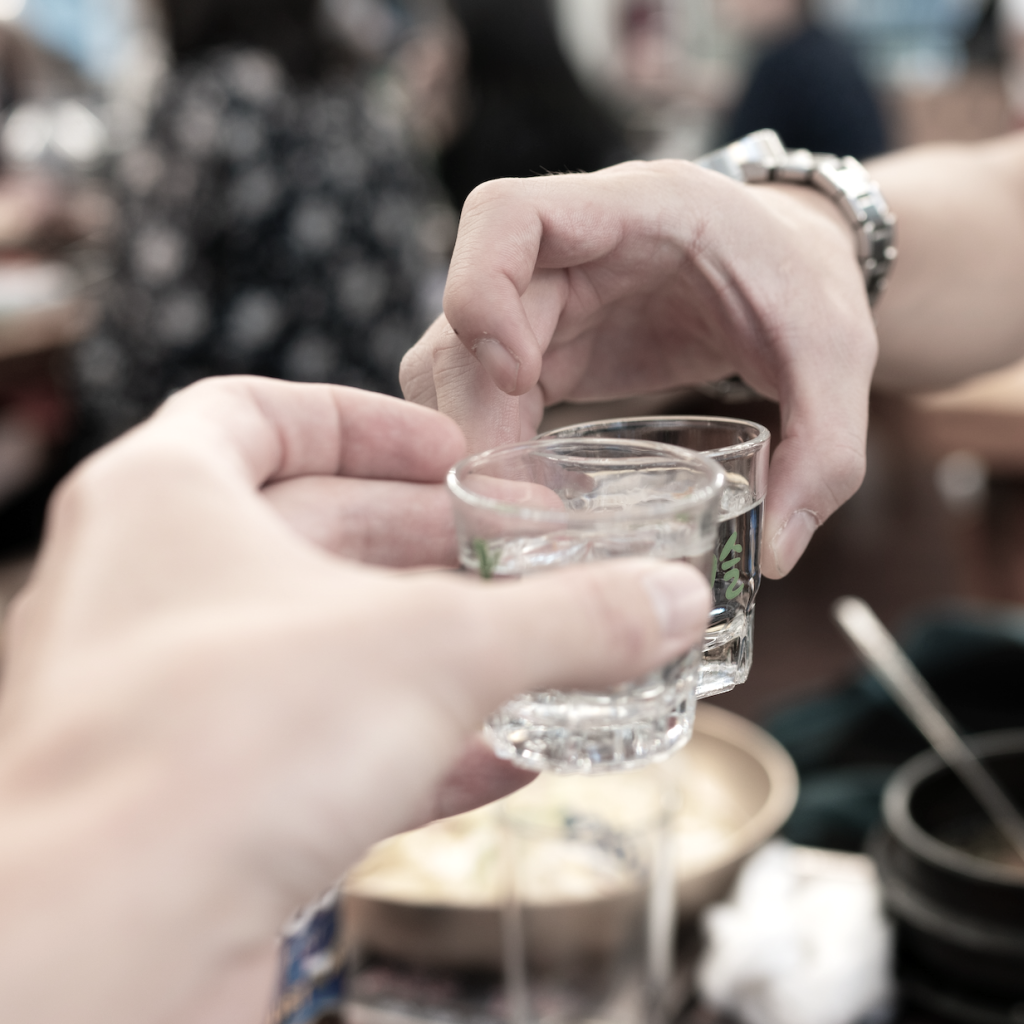 Two hands toasting with shot glasses, sharing a moment with Andong Jinmaek Soju at a lively gathering.