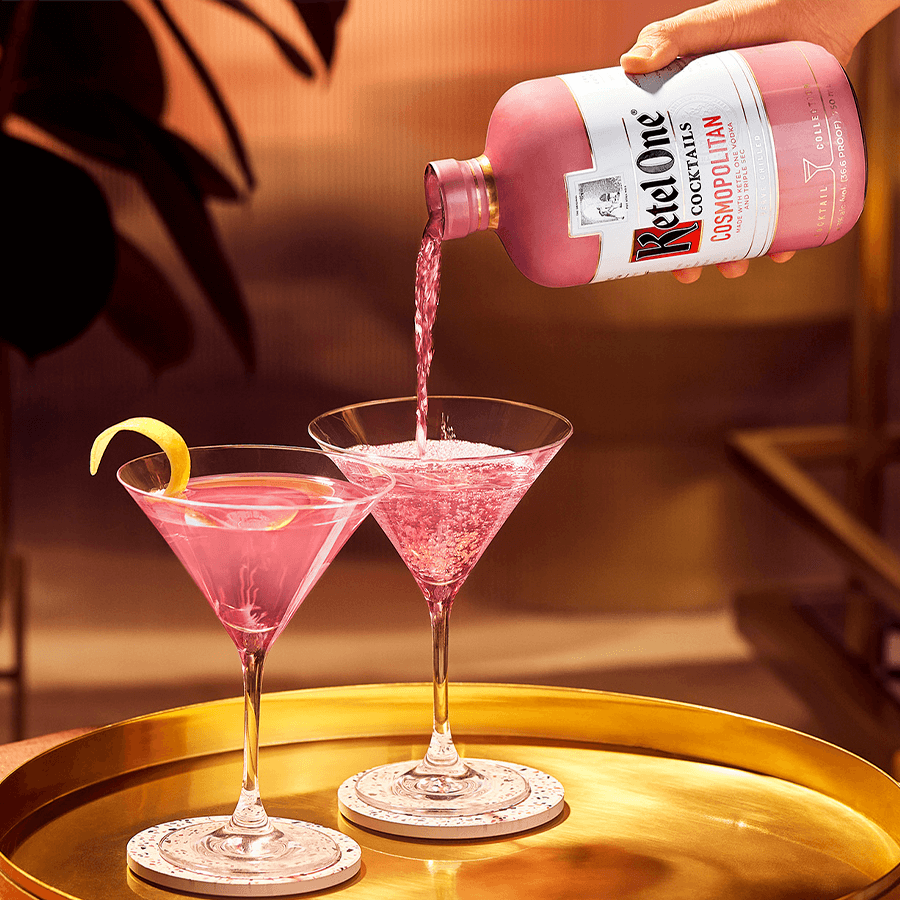 Ketel One Cosmopolitan cocktail being poured into glasses with lemon twist garnish, showcasing its vibrant pink color.