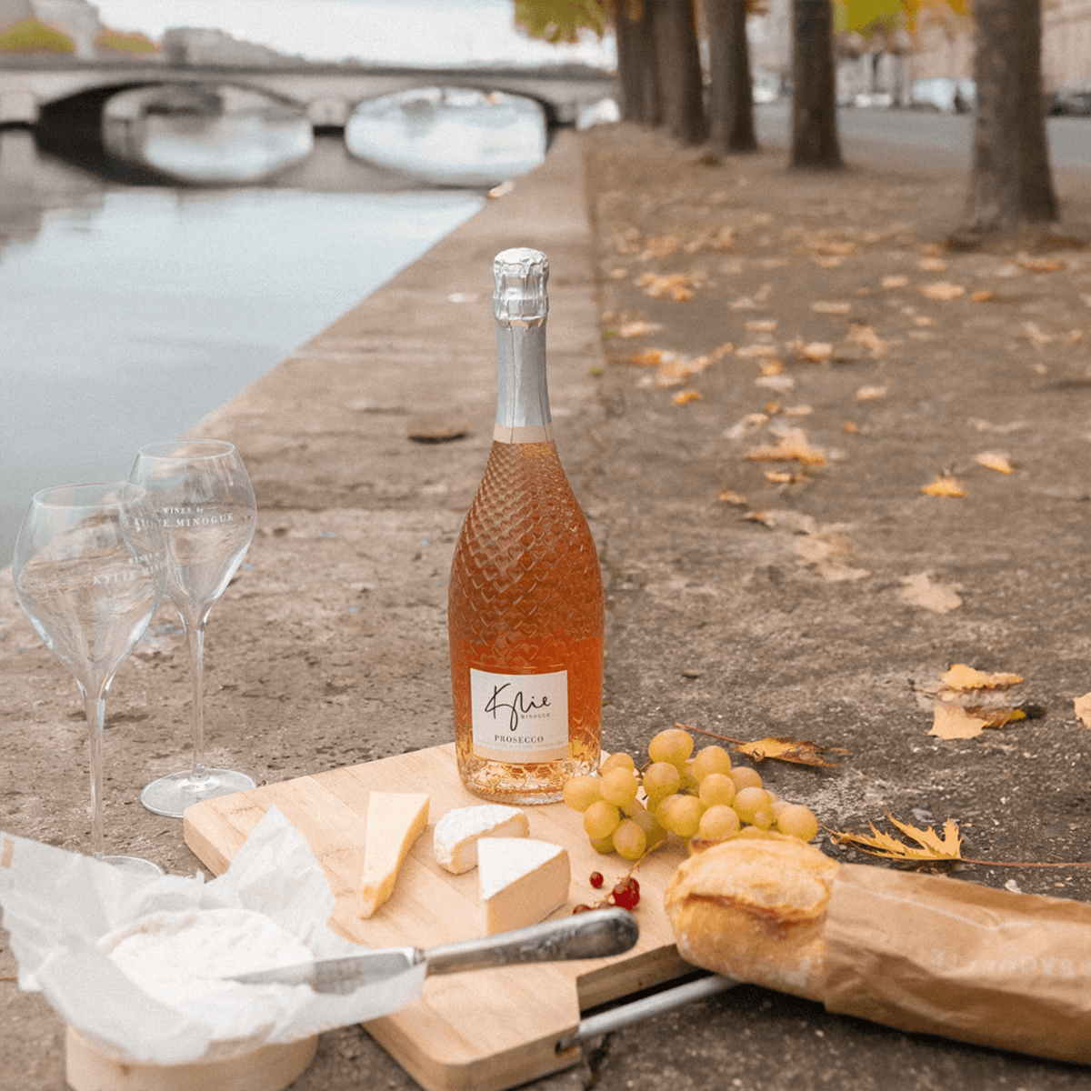 Kylie Minogue Prosecco Rosé bottle with glasses, cheese, and grapes by the riverbank, showcasing a perfect picnic setup.