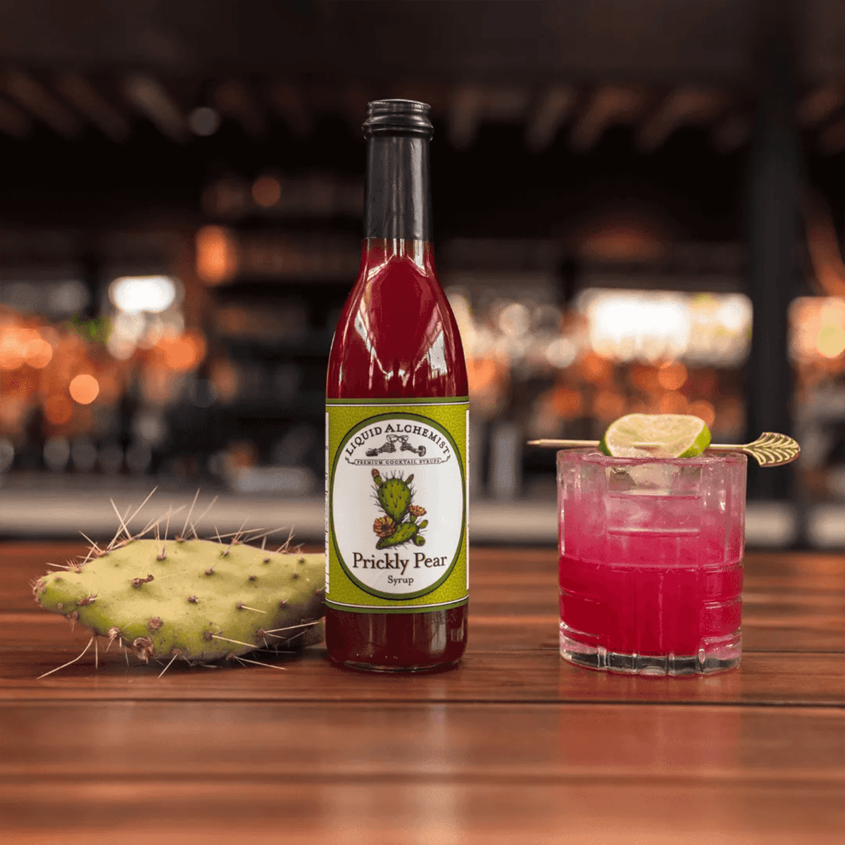 Liquid Alchemist Prickly Pear Syrup bottle near a prickly pear cactus and a vibrant cocktail on a wooden table.