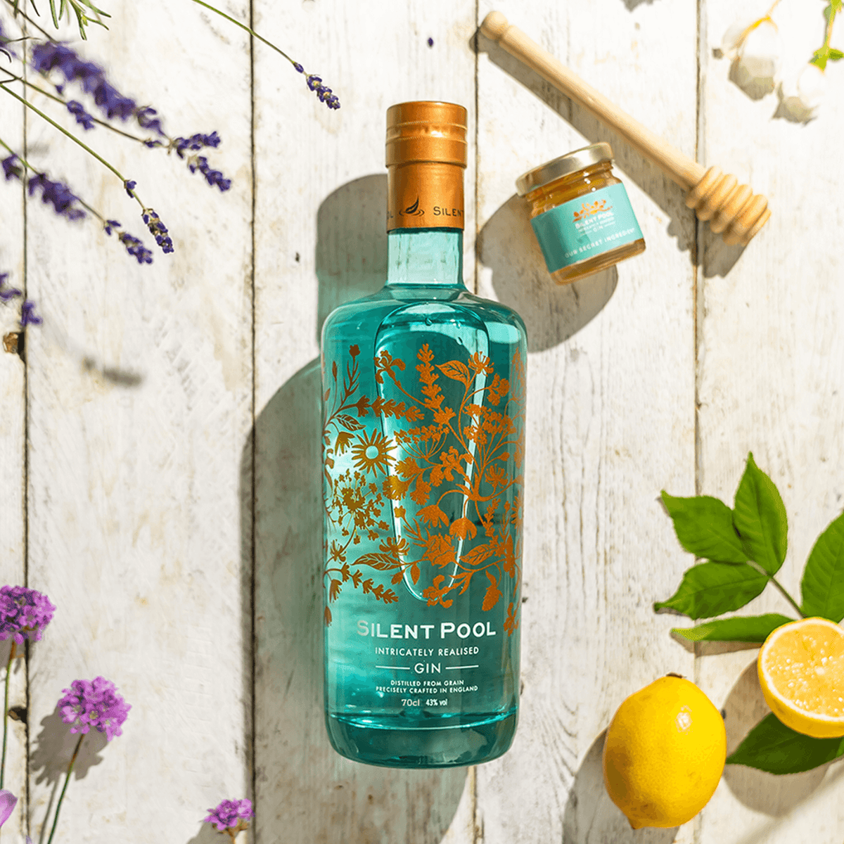Silent Pool Gin bottle with floral design, alongside honey jar and lemon on wooden surface.
