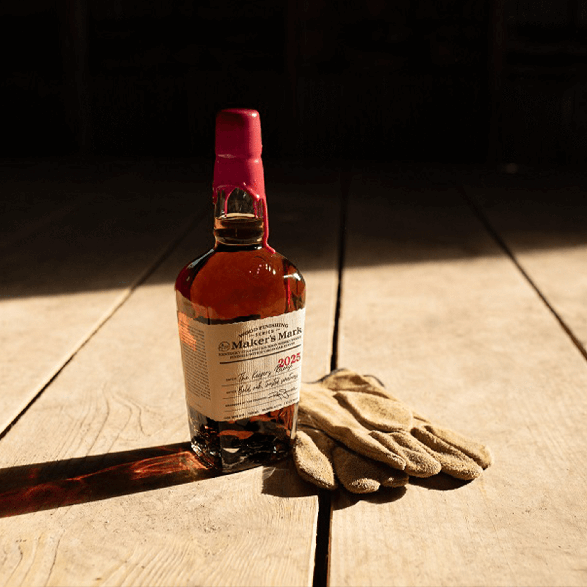 Maker's Mark The Keepers Release 2025 Bourbon bottle with gloves on a wooden surface, highlighting craftsmanship.