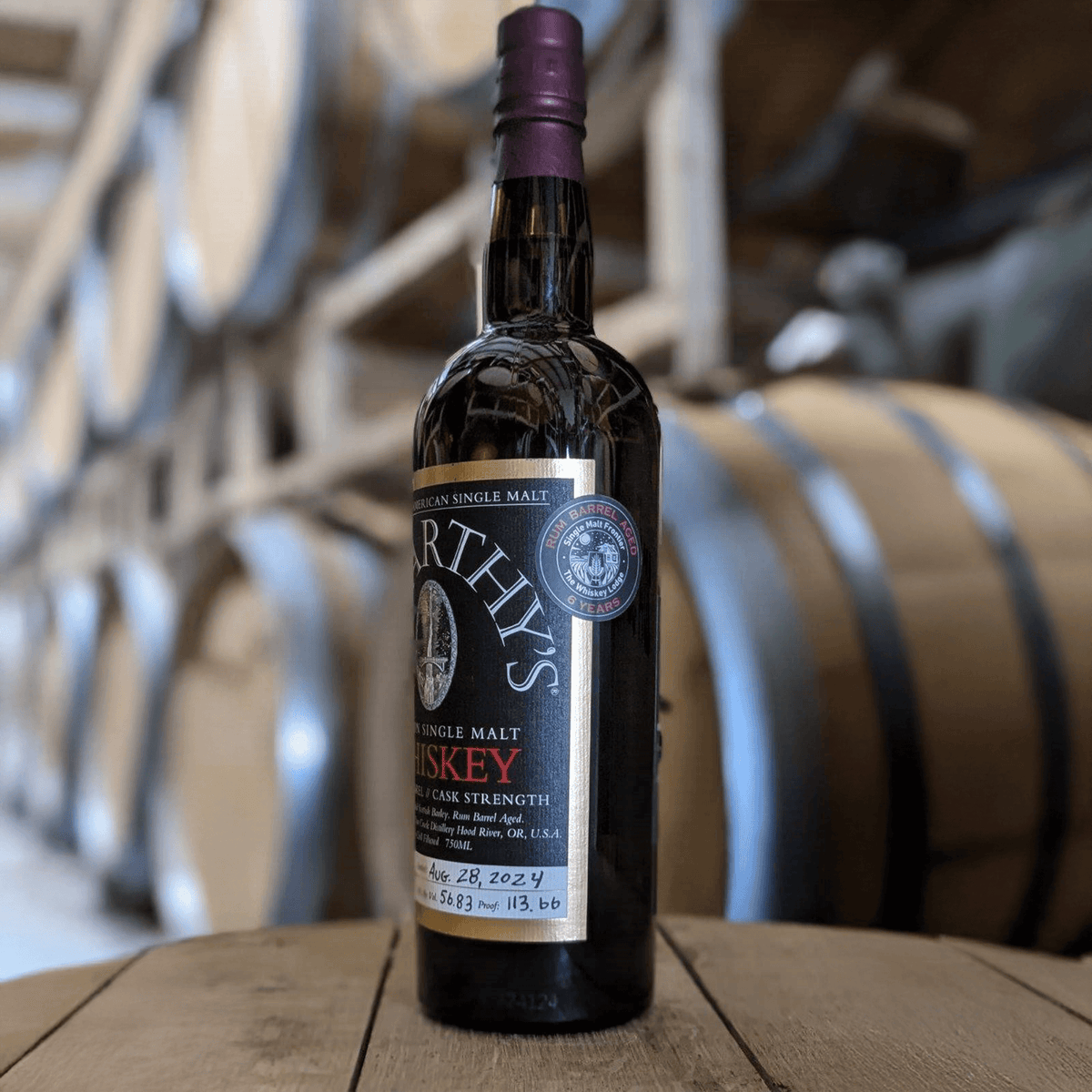 McCarthy’s Single Barrel Cask Strength 6 Year Single Malt Whiskey on a wooden table with barrels in the background.