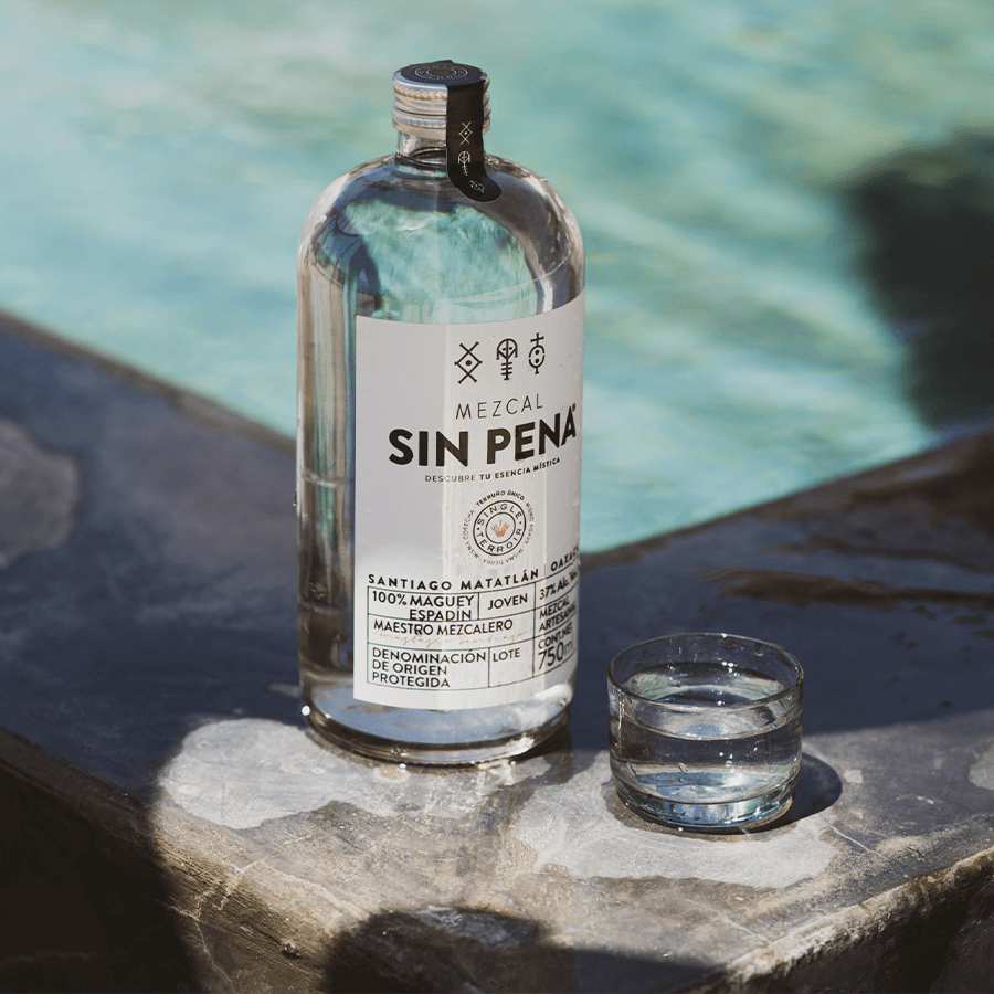 Bottle of Mezcal Sin Pena Joven beside a glass of mezcal, showcasing its artisanal quality by a pool.