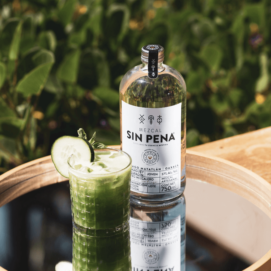 Bottle of Mezcal Sin Pena Joven with a refreshing cocktail on a mirrored table in a natural setting.