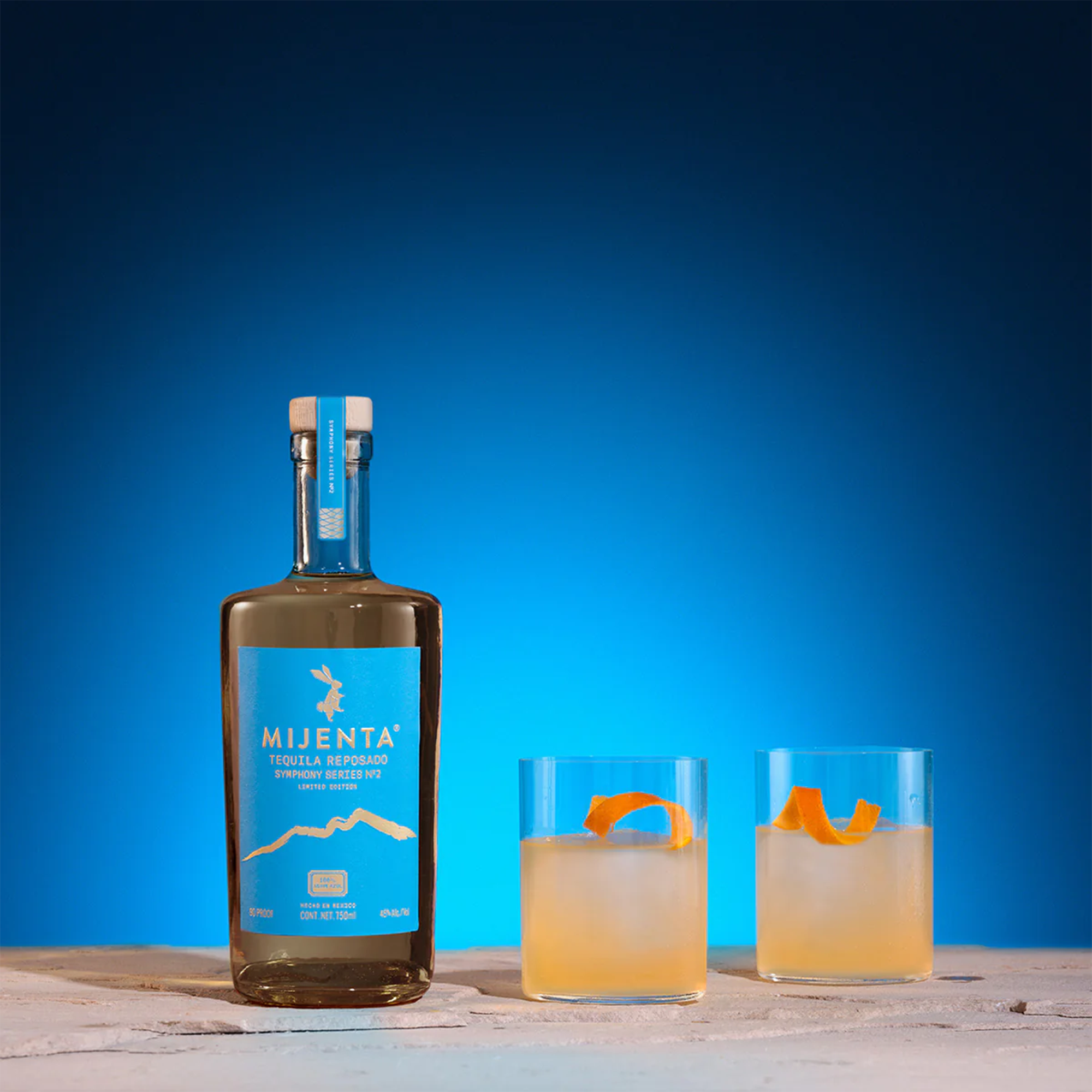 Mijenta Reposado Symphony Series No. 2 Tequila bottle with two cocktails on a blue background.