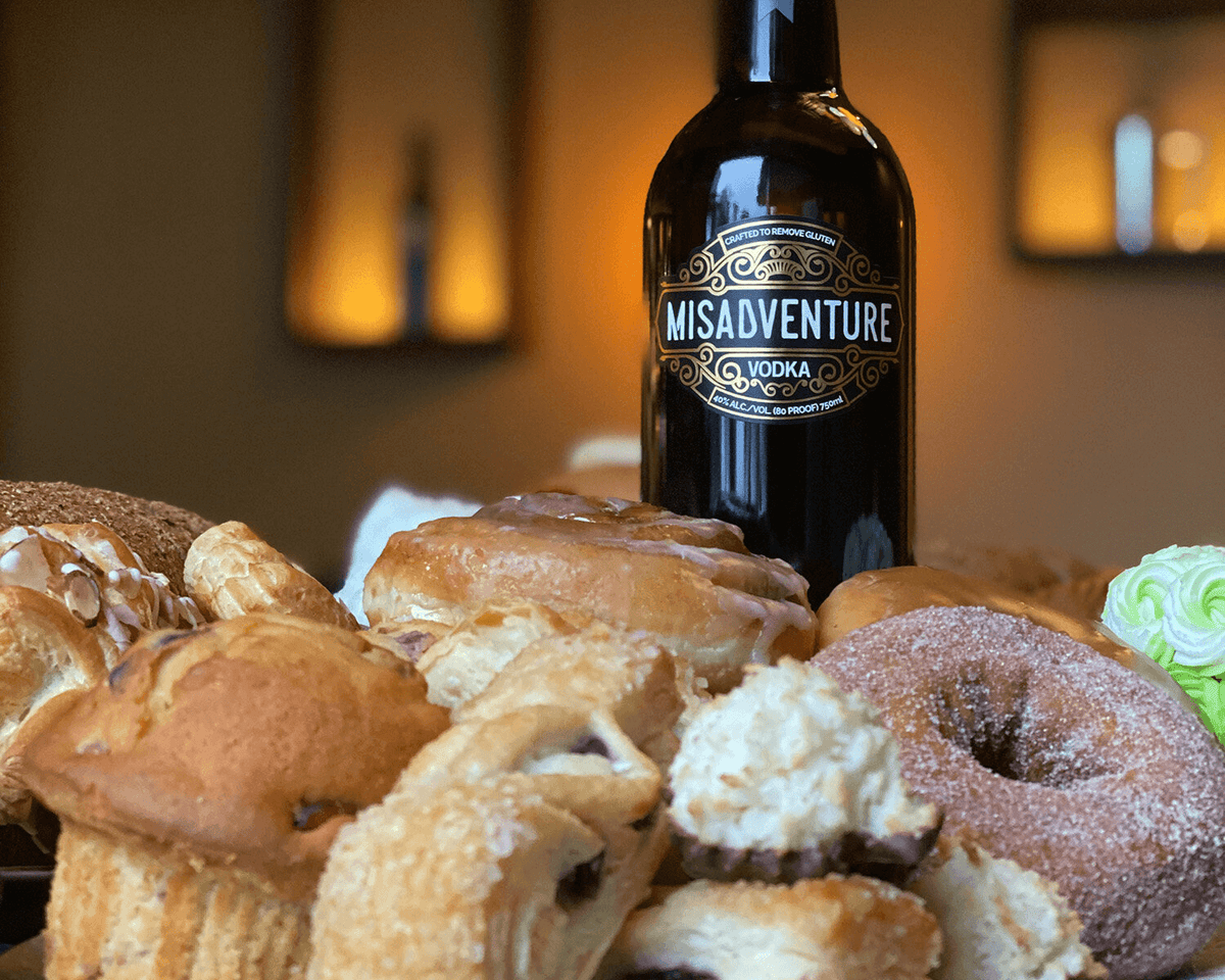 Bottle of Misadventure Sustainable Vodka surrounded by delicious baked goods, symbolizing food waste reduction.