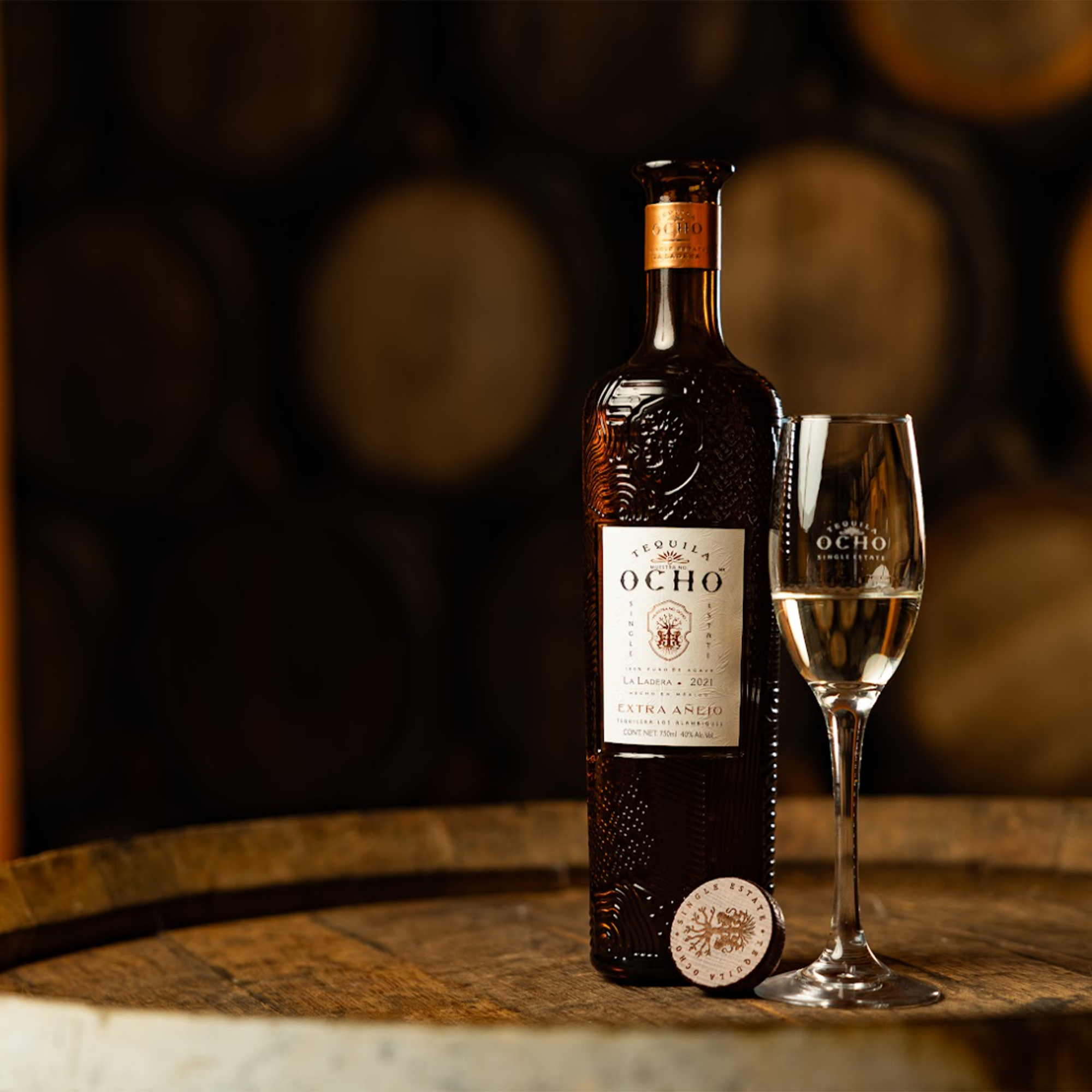 Tequila Ocho La Ladera Extra Añejo bottle and glass on a wooden barrel backdrop, showcasing rich amber color.