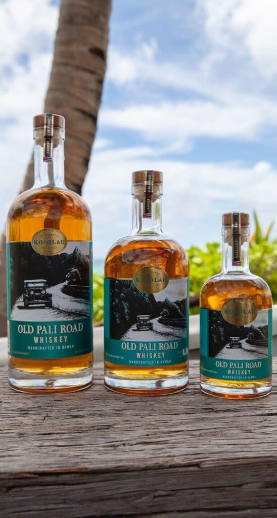 Three bottles of Ko'olau Distillery Old Pali Road Whiskey on a wooden table with a tropical background.