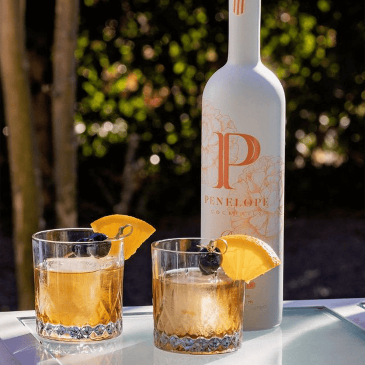 Penelope Peach Old Fashioned cocktails served with oranges and blueberries, showcasing the elegant bottle outdoors.