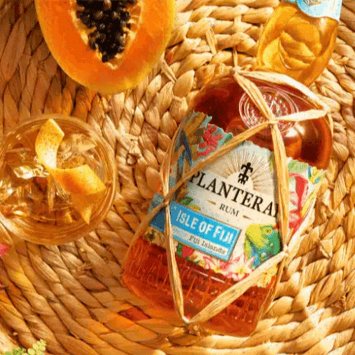 Planteray Double Barrel Isle of Fiji Rum bottle on woven mat, surrounded by tropical fruits and a cocktail.