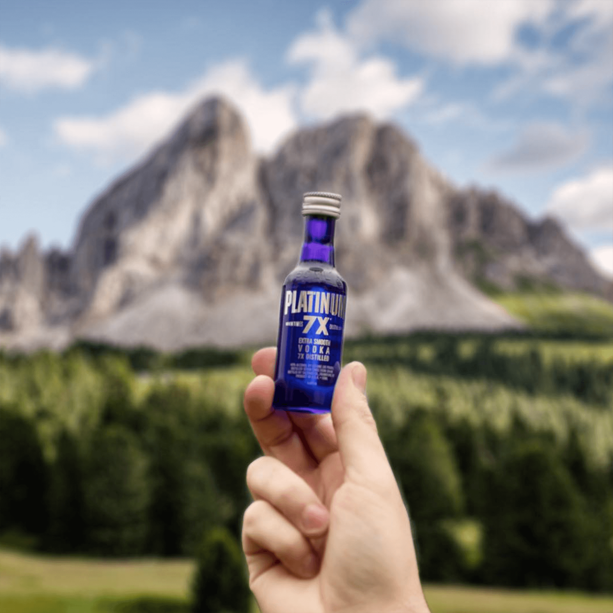 Hand holding a 50ml bottle of Platinum 7x Vodka against a mountain backdrop, showcasing its striking blue design.