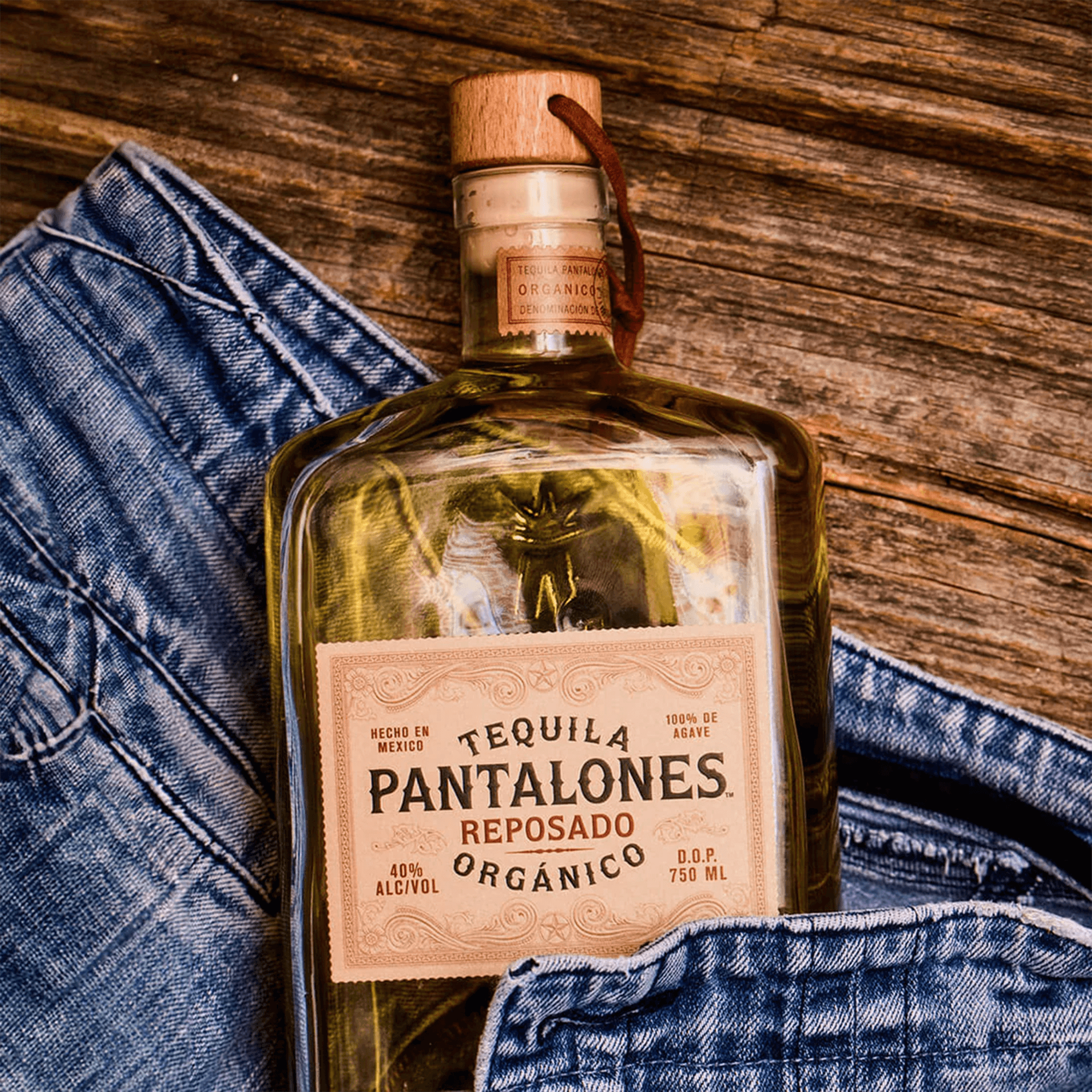 Pantalones Reposado Tequila bottle on a wooden table next to denim jeans, showcasing its organic agave and unique aging.