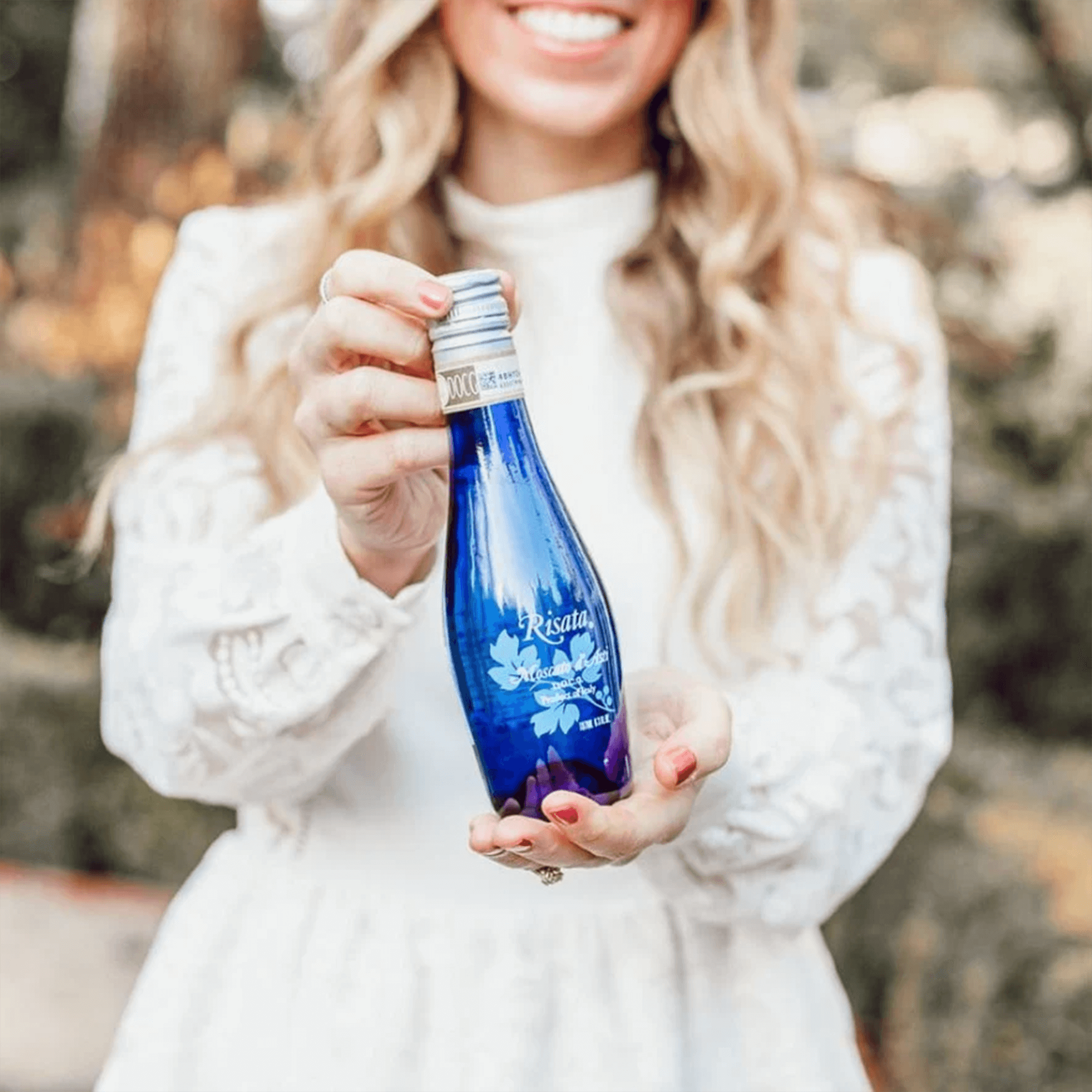Woman holding a bottle of Risata Moscato d'Asti 187ml, showcasing its vibrant blue design and label in a bright outdoor setting.