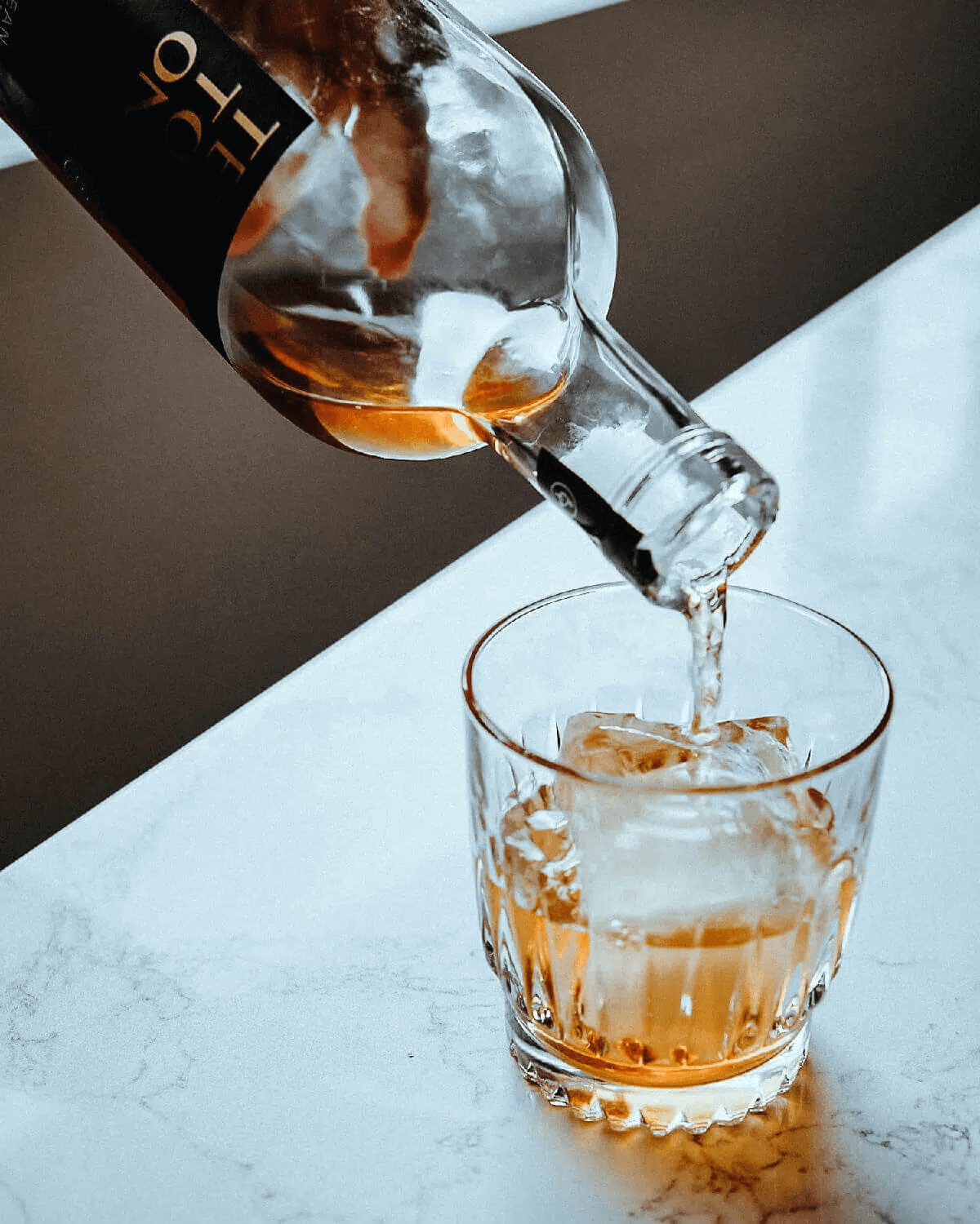 Pouring Ten To One Caribbean Dark Rum over ice in a glass on a marble surface.
