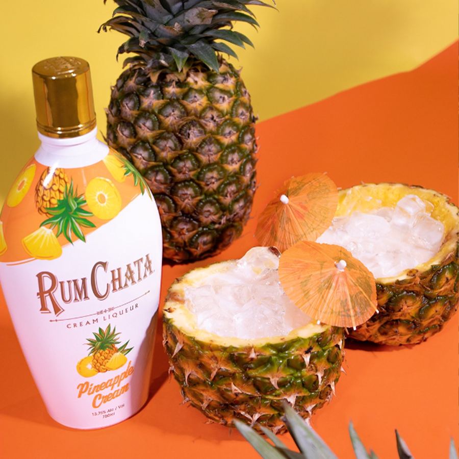 RumChata Pineapple bottle surrounded by fresh pineapples and tiki glasses, perfect for tropical cocktails.