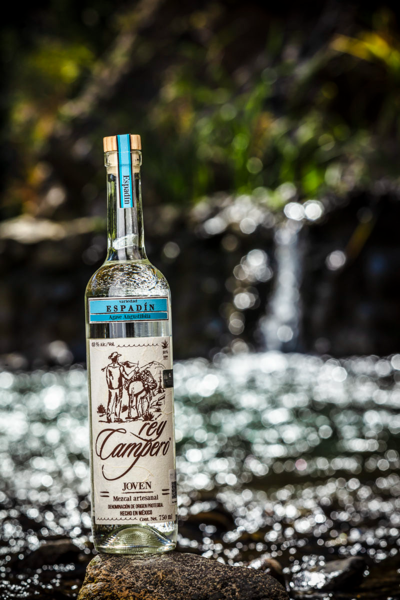 Bottle of Rey Campero Espadin Mezcal displayed outdoors with a blurred waterfall in the background.