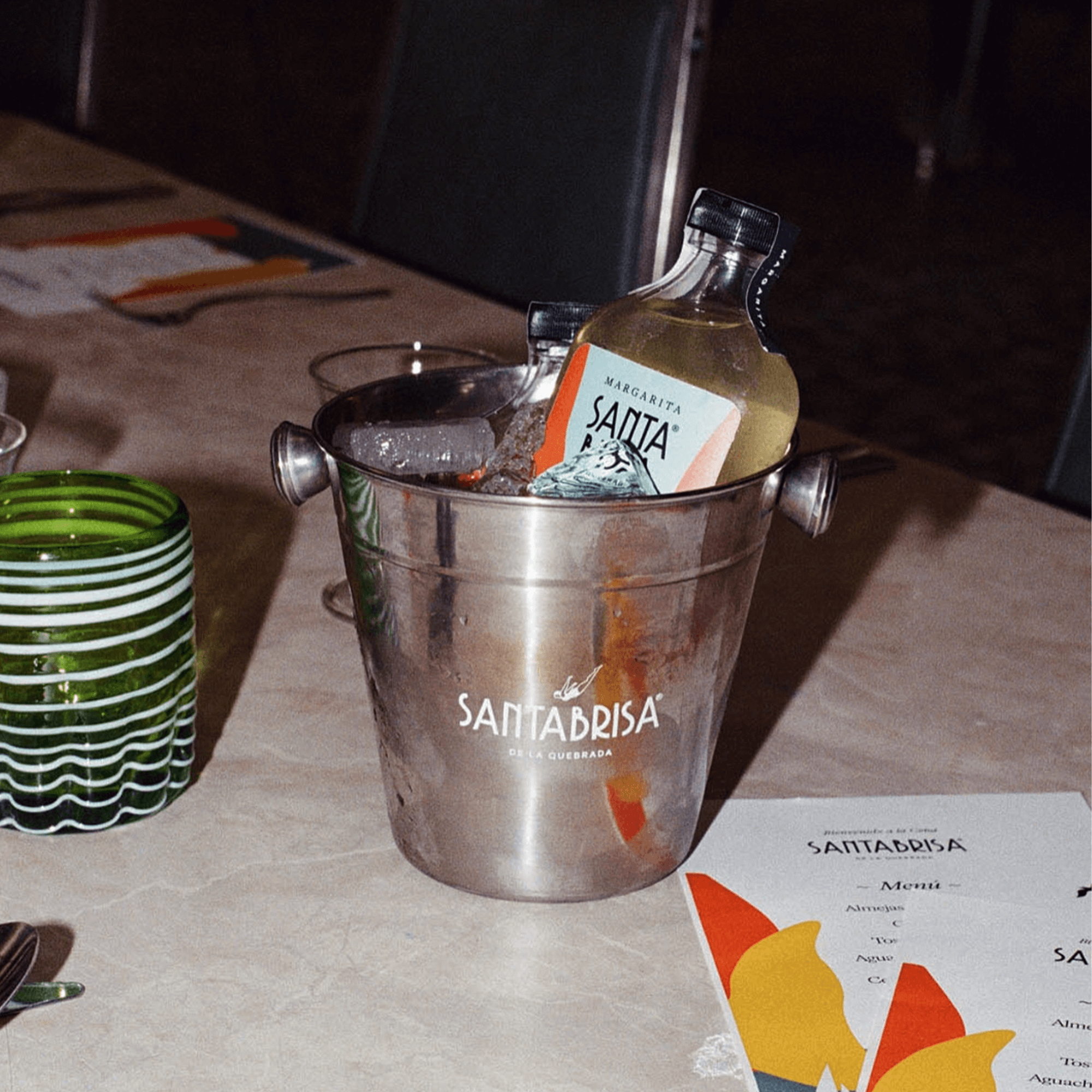 Santa Brisa margarita in a bucket with ice, featured on a table alongside colorful glasses and a menu.
