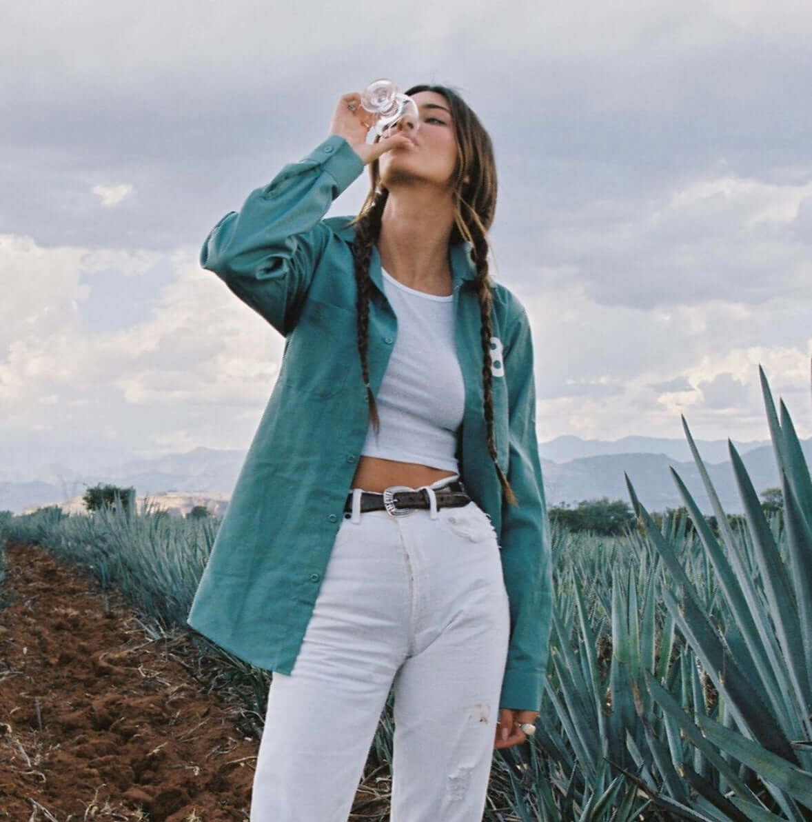 Woman enjoying tequila in a blue shirt, standing in a lush agave field under a cloudy sky.