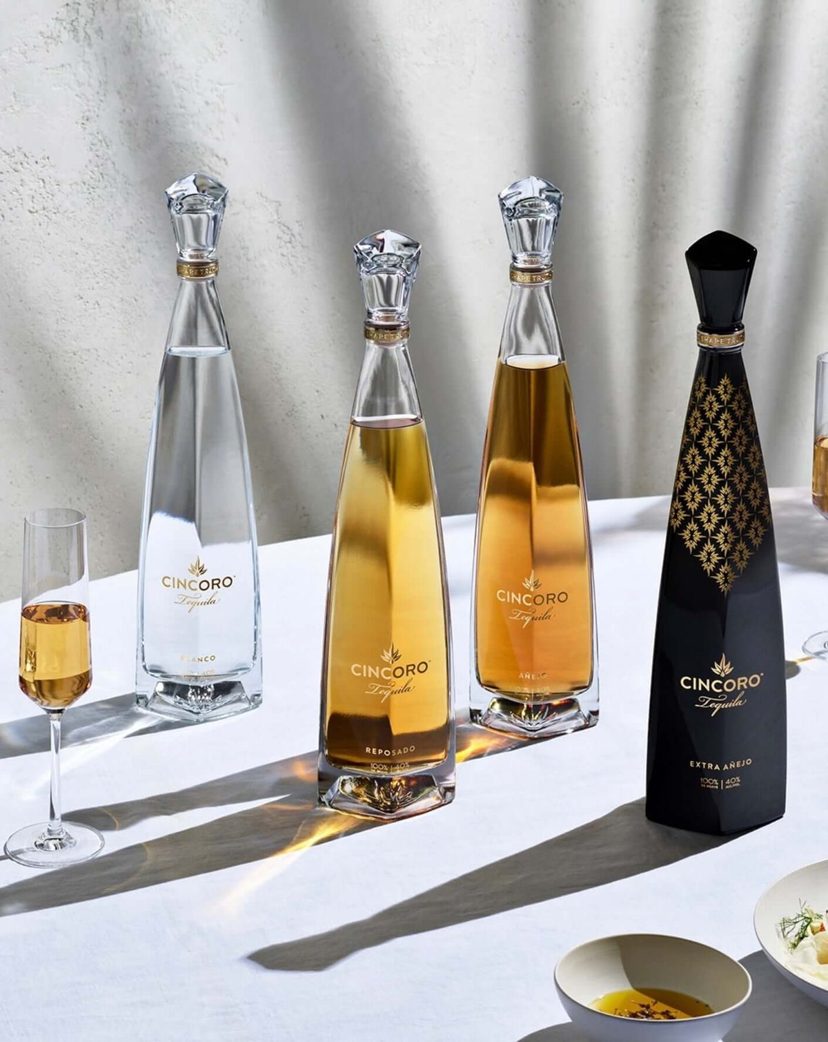 Cincoro Tequila bottles including Blanco, Reposado, and Extra Añejo on a table with glasses and small plates.