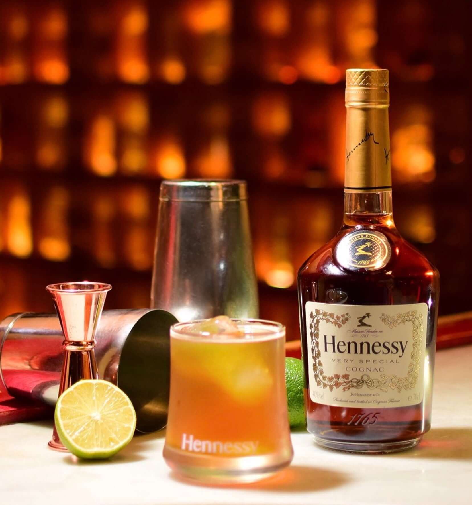 Hennessy Very Special V.S Cognac bottle with cocktail, lime, and bar tools on a warm bar background.