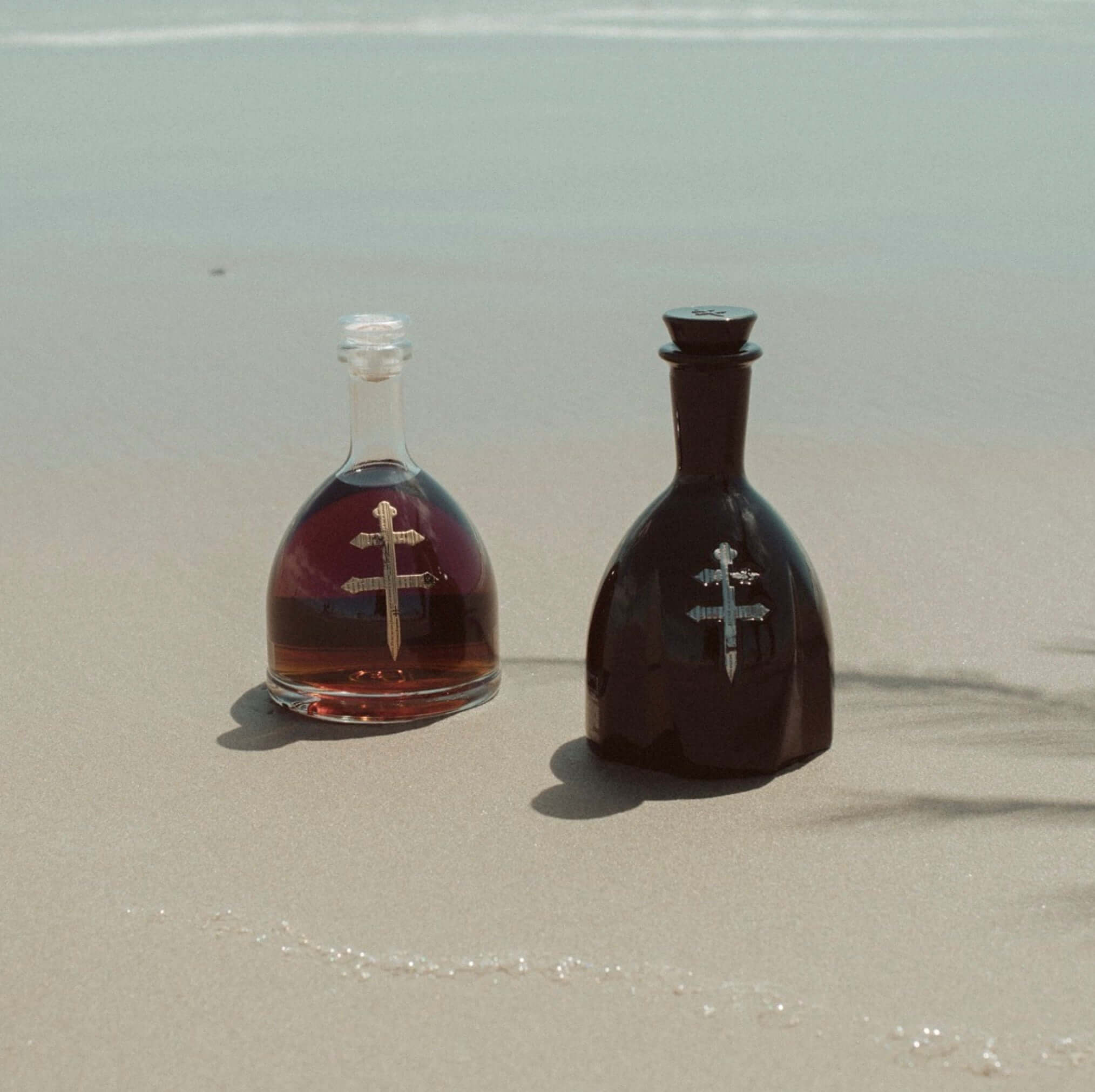 Two D'USSÉ VSOP Cognac bottles on the beach, showcasing rich amber colors against a sandy backdrop.