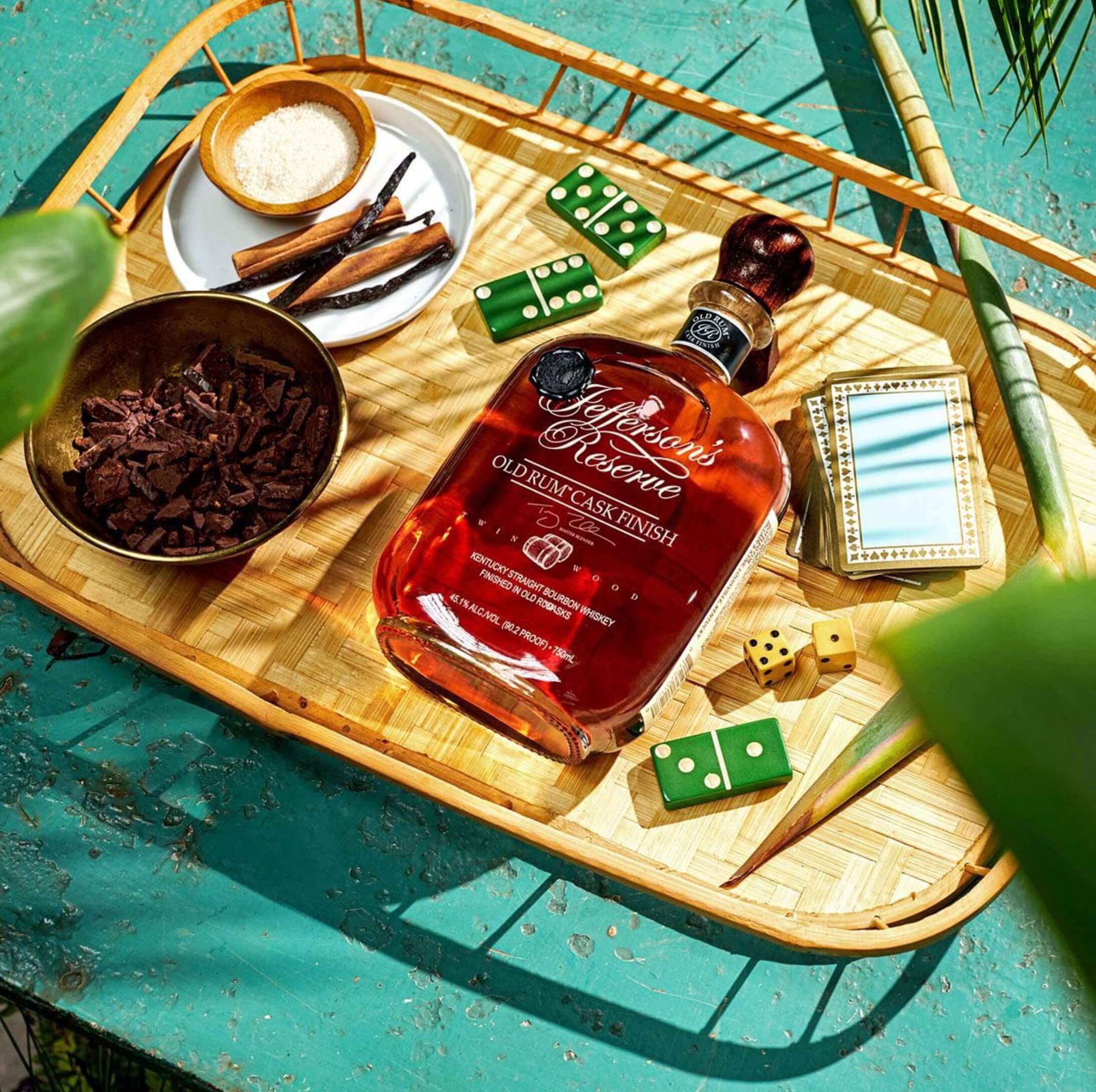 Jefferson's Reserve Old Rum Cask Finish Bourbon bottle on a bamboo tray with spices and games, evoking a tropical vibe.
