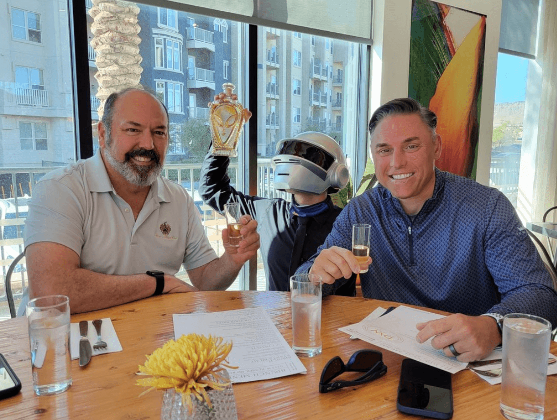 Two men at a restaurant holding tequila shots with a robot figure, celebrating with Alien Añejo Tequila.
