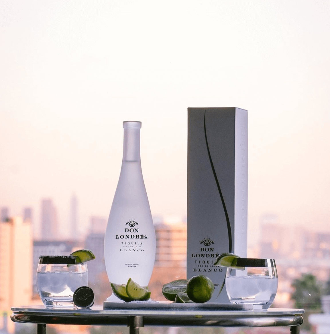 Don Londrés Blanco Tequila bottle with citrus garnish and glasses against a city skyline background.