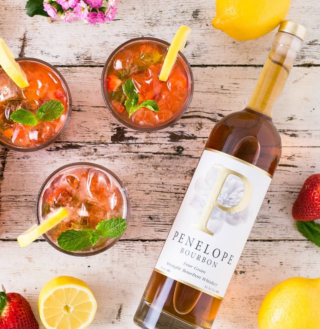 Penelope Four Grain Straight Bourbon bottle with cocktails, lemon, and strawberries, perfect for summer sipping.