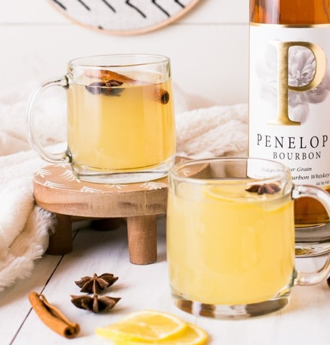 Penelope Four Grain Straight Bourbon served in two glasses with spices and lemon, on a cozy backdrop.