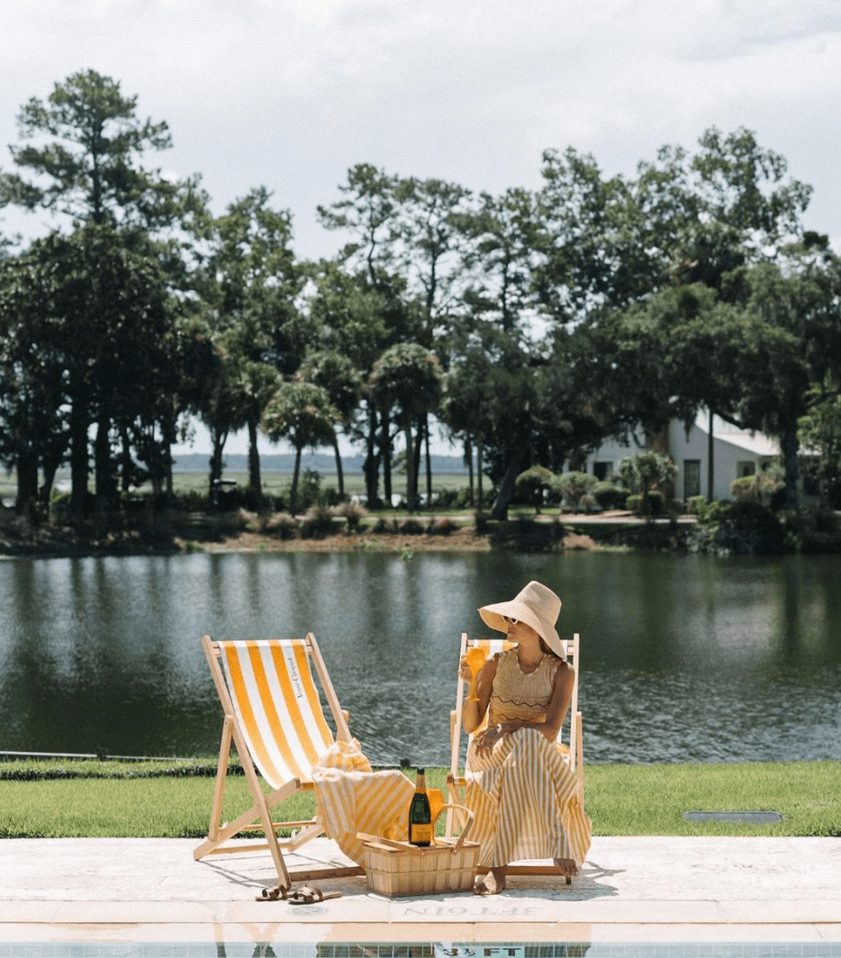 Woman in a sunhat relaxing by a pool with Veuve Clicquot Champagne and picnic basket in a serene outdoor setting.