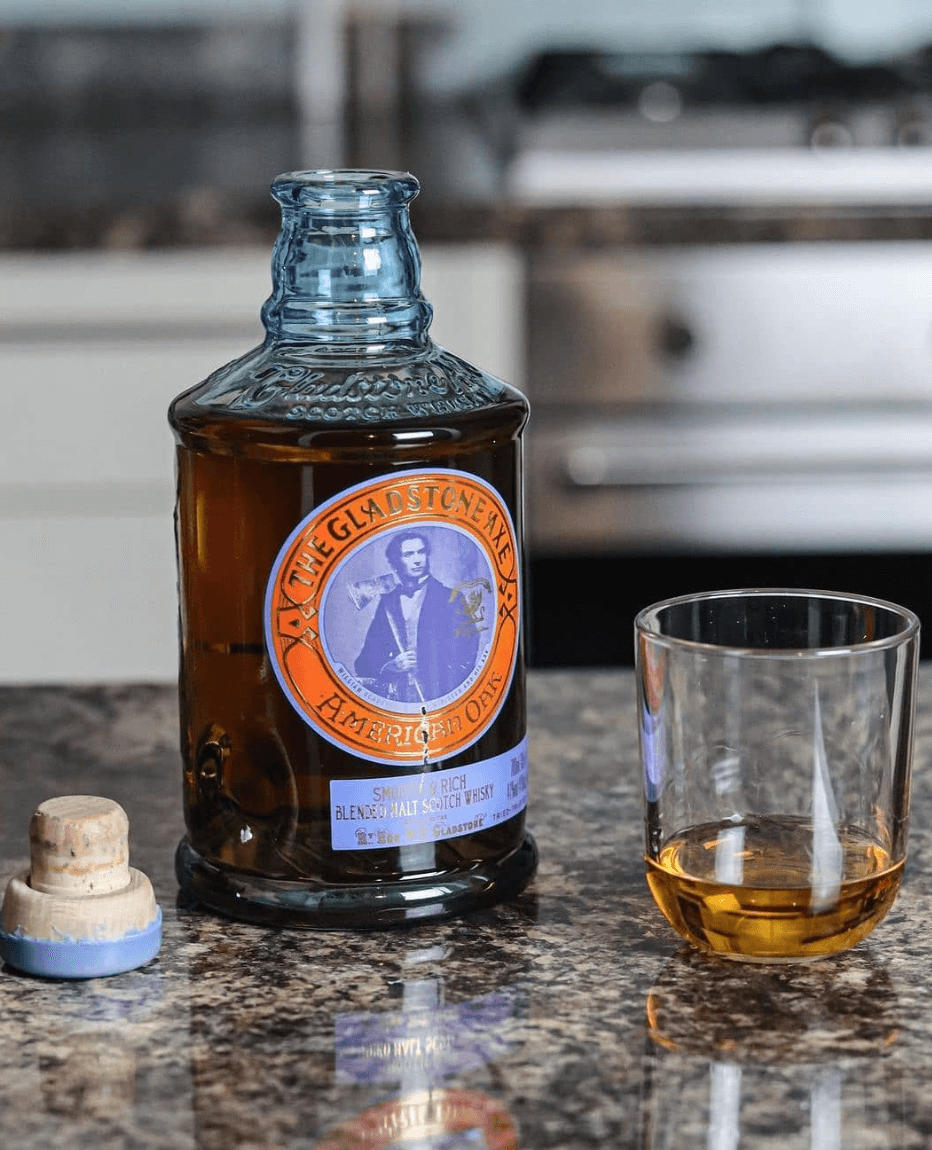 Gladstone Axe American Oak blended scotch whisky bottle with a glass, showcasing rich amber color on a granite countertop.