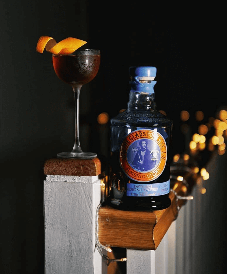 Gladstone Axe American Oak Scotch bottle with a cocktail and orange peel garnish, set against a dark background.
