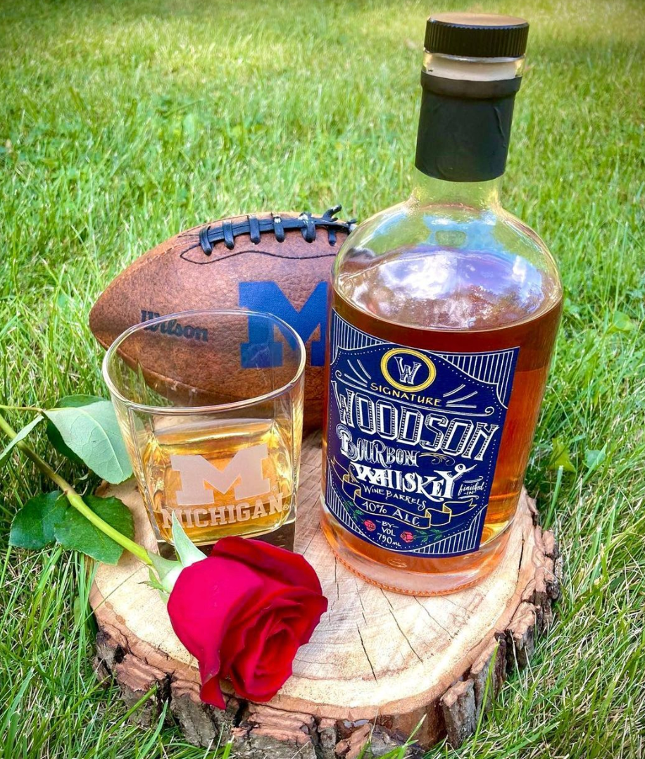 Woodson Whiskey Blue & Maize Bourbon bottle with a glass, rose, and football on a wooden stump outdoors.