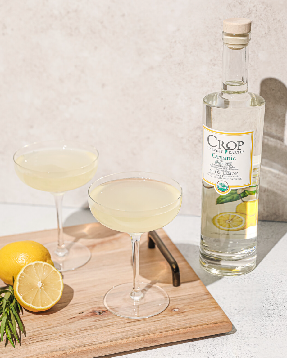 Crop Organic Vodka bottle beside two lemon cocktails and fresh lemons on a wooden cutting board.