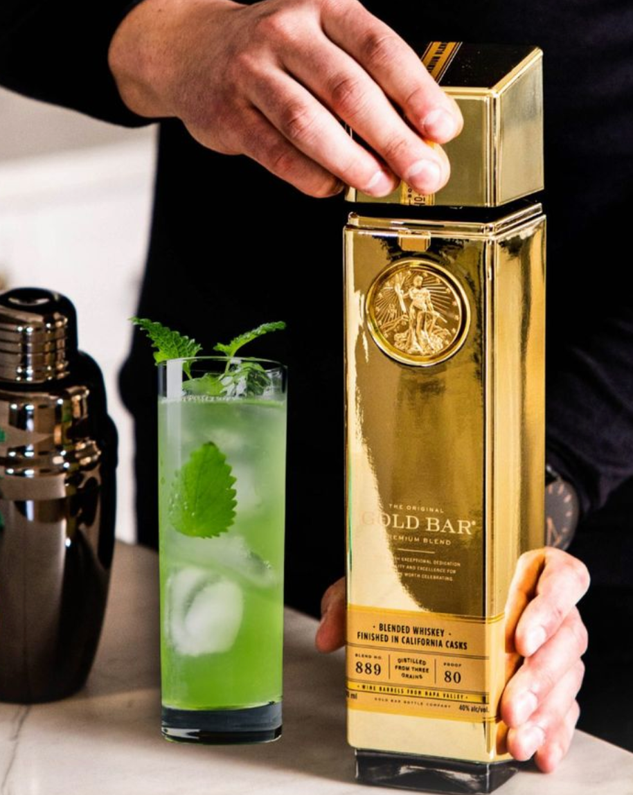 Hand holding a bottle of Gold Bar Original Premium Blended Whiskey next to a refreshing cocktail with mint leaves.