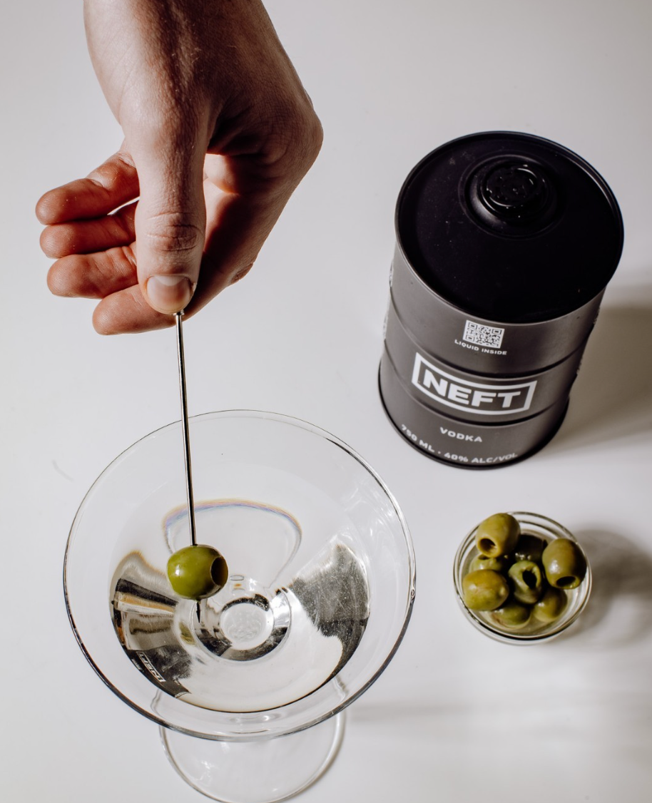 Hand selecting an olive for a cocktail with NEFT Black Barrel Australian Vodka canister and olives on a table.