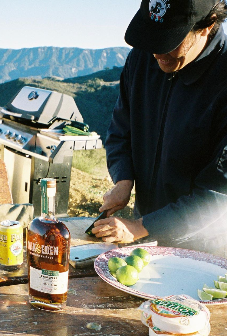 Outdoors scene with Oak & Eden Rye & Spire Whiskey bottle, limes, and grill in the background.