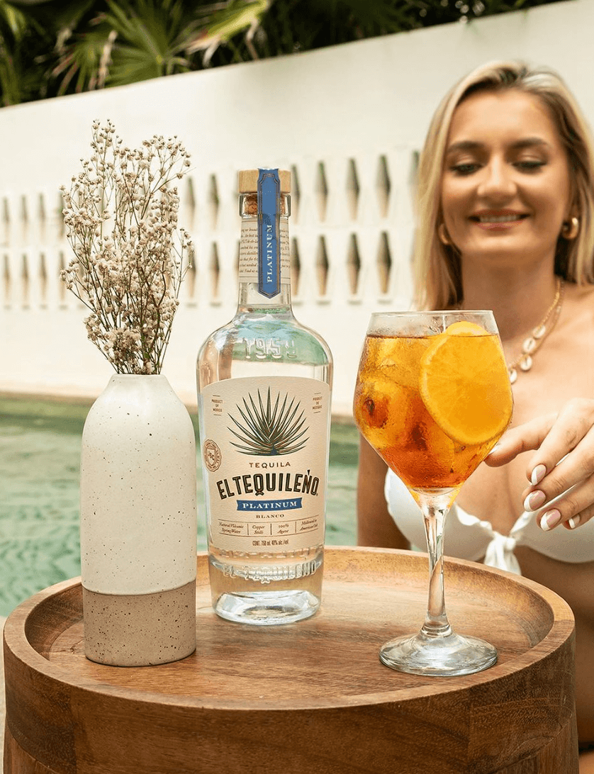 El Tequileño Platinum Blanco Tequila bottle with cocktail and decorative vase on a wooden table by a poolside.